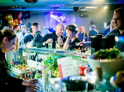 Lebhafter Hotelbar-Abend am beleuchteten Tresen, Barkeeper mixt Cocktails, Gäste im Gespräch, Disco-Lichter und Obstgarnitur