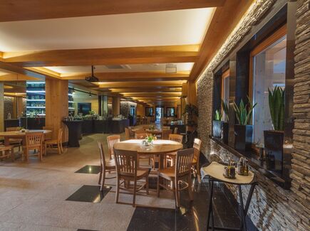 Gemütliches Hotelrestaurant mit sichtbaren Holzbalken, steinerner Akzentwand, runden Holztischen, Bar- und Buffetbereich, Pflanzen auf der Fensterbank