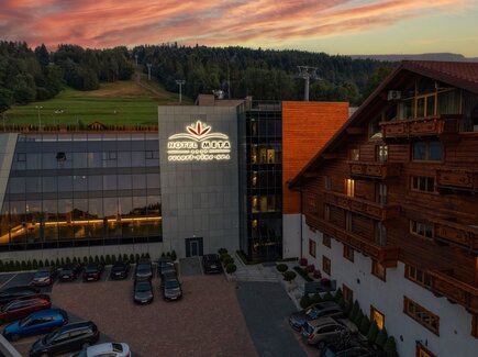 Hotel Meta Resort & Spa mit leuchtendem Logo, Glasfassade mit Blick auf Hallenbad, angrenzender Holzchalet-Flügel und bewaldete Hänge bei Sonnenuntergang