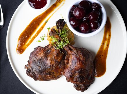 Zart geschmorte Entenkeule mit brauner Sauce, Kräutern, Kartoffelplätzchen und glasierten Kirschen in Schale auf weißem Teller