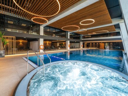 Überdachtes Hotel-Spa mit sprudelndem Whirlpool im Vordergrund, langem Schwimmbecken, Liegen, Holzlamellendecke und runden Hängeleuchten.