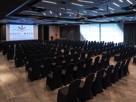 Konferenzsaal im Theaterstil mit Reihen schwarzer Stuhlhussen, großer Projektionsleinwand mit Hotel-Logo, Rednerpult und schlichten Vorhängen