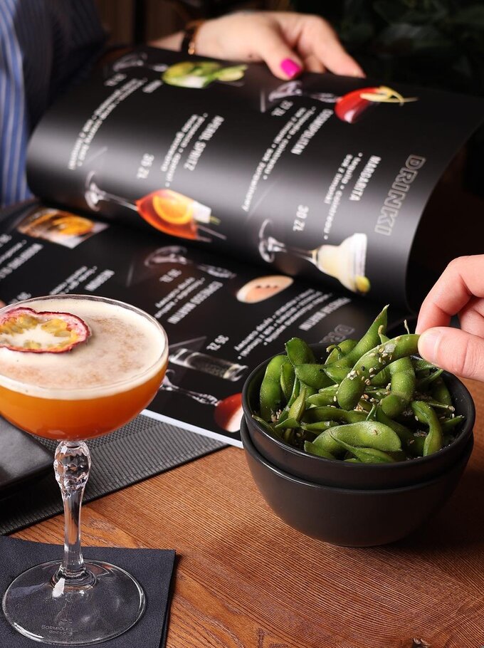 Passionsfrucht-Cocktail und Edamame mit Sesam auf Holztisch, Gast blättert in der Getränkekarte und greift eine Schote