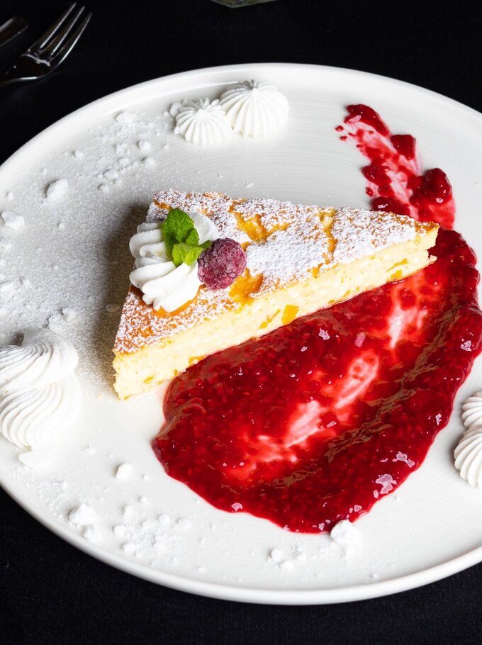 Stück Käsekuchen mit Puderzucker, Sahne, Himbeere und Minze, serviert mit Himbeercoulis und Baiser-Tupfen auf weißem Teller