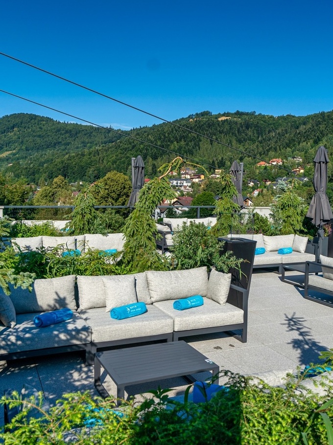 Rooftop lounge with gray cushioned sofas, blue rolled towels, closed parasols and potted evergreens, overlooking a green mountain village