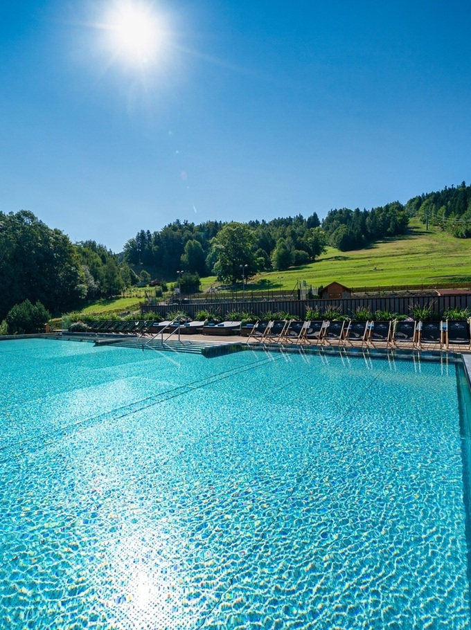 Outdoor pool with sunbeds and rolled towels on a wooden deck, view of sunlit mountain slopes, forest and chairlift