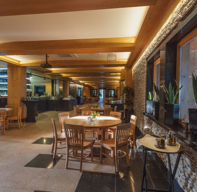 Warm hotel dining room with exposed wooden beams, stone accent wall, round wooden tables, bar and buffet area, potted plants on the windowsill