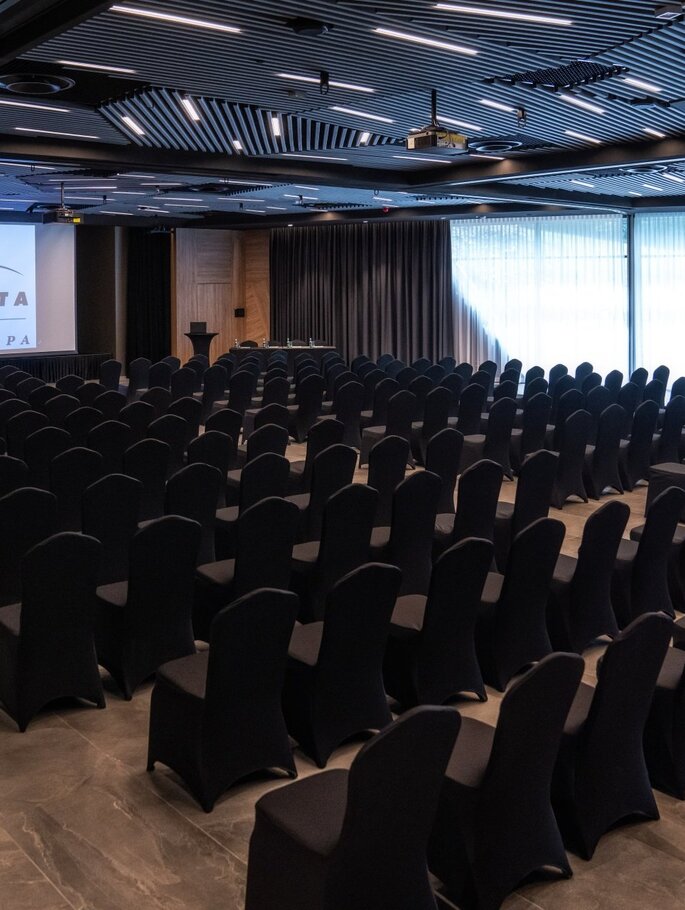 Konferenzsaal im Theaterstil mit Reihen schwarzer Stuhlhussen, großer Projektionsleinwand mit Hotel-Logo, Rednerpult und schlichten Vorhängen