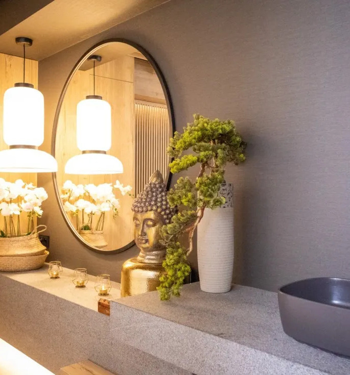 Spa-ähnliche Waschtischzone mit rundem Spiegel, warmen Hängeleuchten, Orchideen, goldener Buddha-Figur, Bonsai, Steinwaschbecken und Messingarmatur.