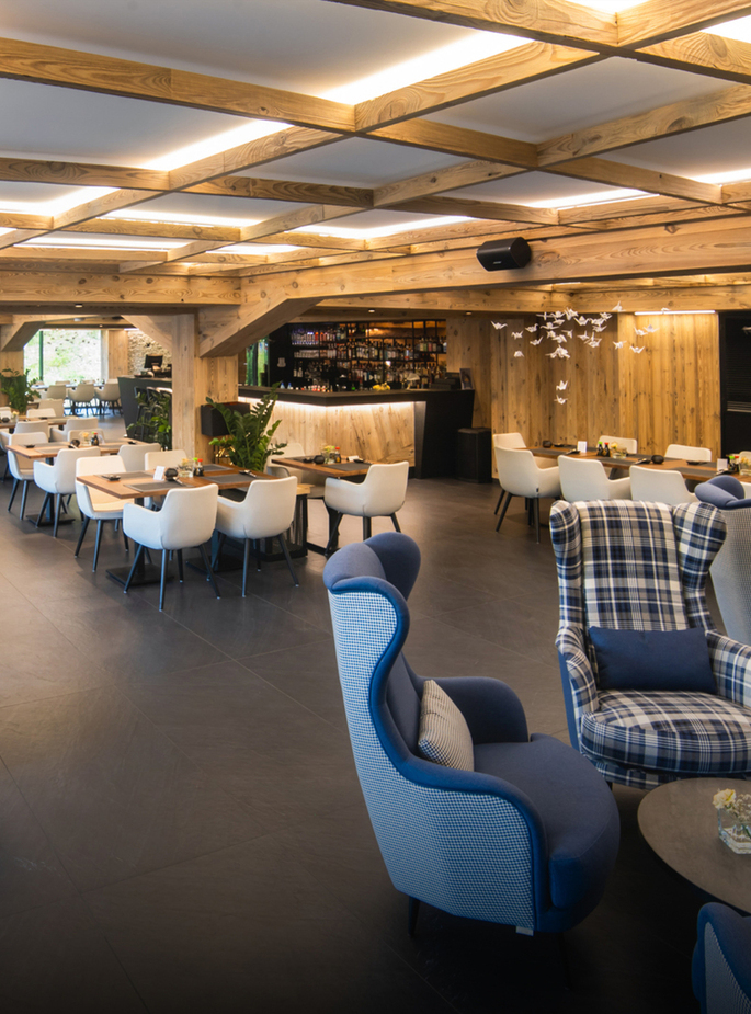 Cozy hotel lounge with wooden-beam ceiling, bar, white dining chairs, blue plaid armchairs, plants and warm lighting.