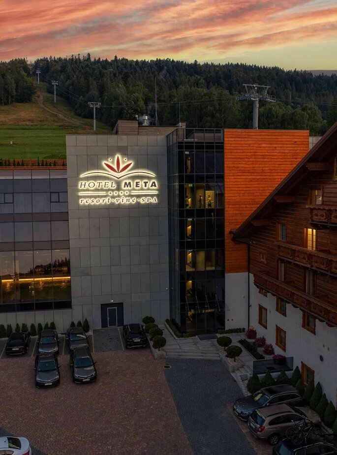 Hotel Meta Resort & Spa mit leuchtendem Logo, Glasfassade mit Blick auf Hallenbad, angrenzender Holzchalet-Flügel und bewaldete Hänge bei Sonnenuntergang