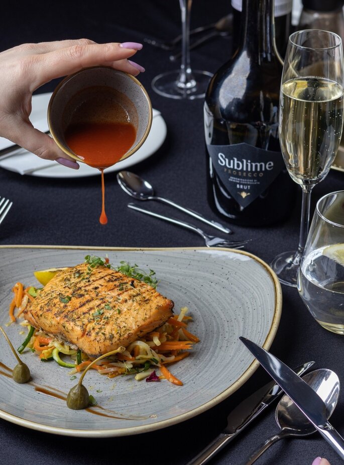 Gegrillter Lachs auf Gemüse mit Kapernbeeren, Hand gießt rote Sauce; Flasche Sublime Prosecco und Sektflöte