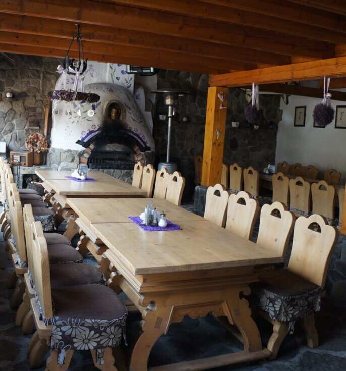 Rustic dining room with long wooden communal tables, cushioned chairs, stone wood-fired oven and purple floral decorations.