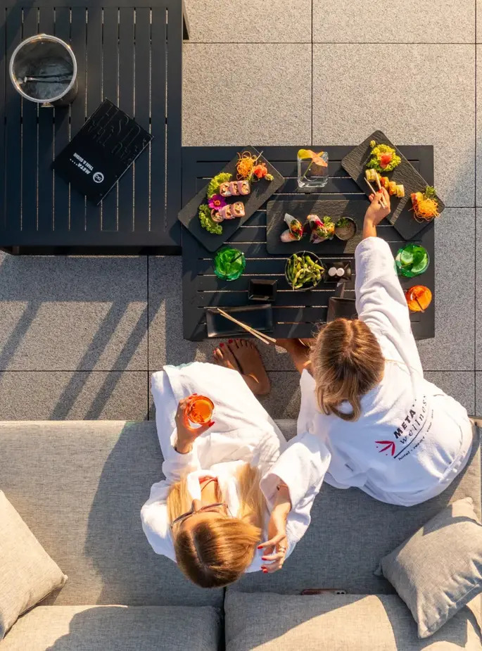 Guests in white spa robes enjoying sushi platters and cocktails on a sunny hotel rooftop lounge with modern tile flooring.