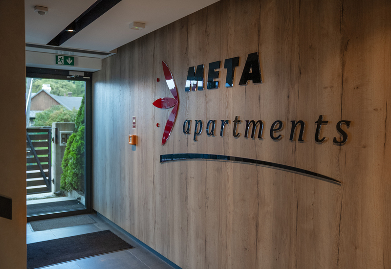 Wood-paneled lobby with META apartments wall sign, glass exit door revealing garden greenery and mats at the modern entrance.