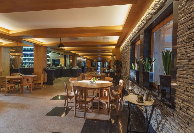 Warm hotel dining room with exposed wooden beams, stone accent wall, round wooden tables, bar and buffet area, potted plants on the windowsill