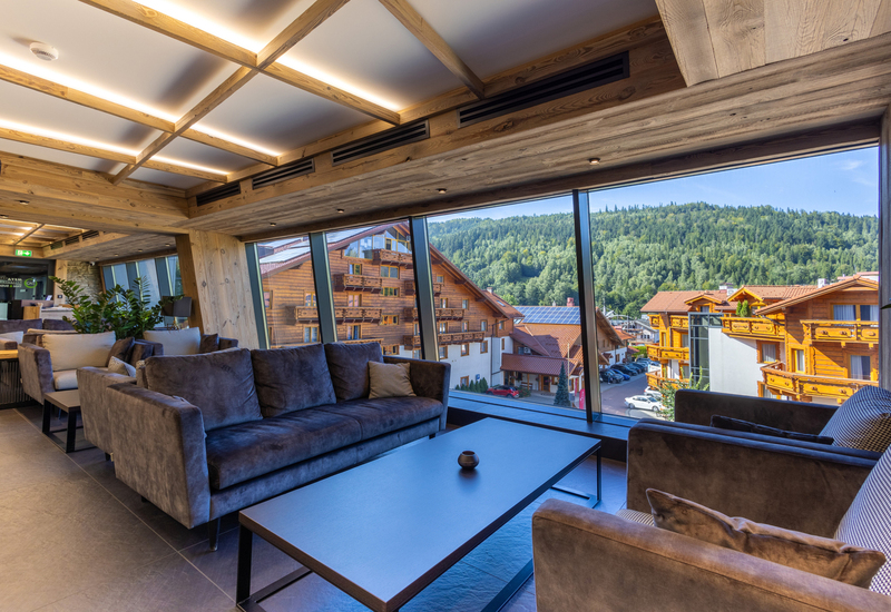 Cozy hotel lounge with plush sofas, wood-beamed ceiling and floor-to-ceiling windows overlooking alpine chalets and forested hillside