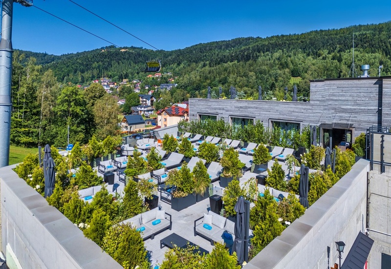 Rooftop hotel terrace with cushioned loungers, potted evergreens, umbrellas and mountain village view with cable car.