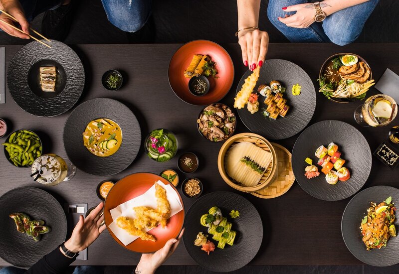 Shared modern Asian tasting spread — sushi, tempura, ramen, edamame on black slate plates, hands reaching to serve.