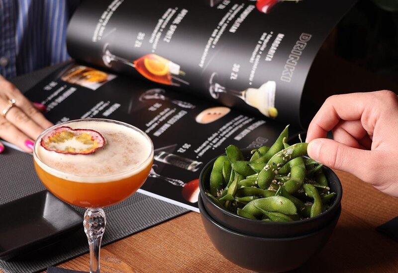 Passion-fruit cocktail and sesame edamame on a wooden table, guest browsing the cocktail menu and reaching for a pod