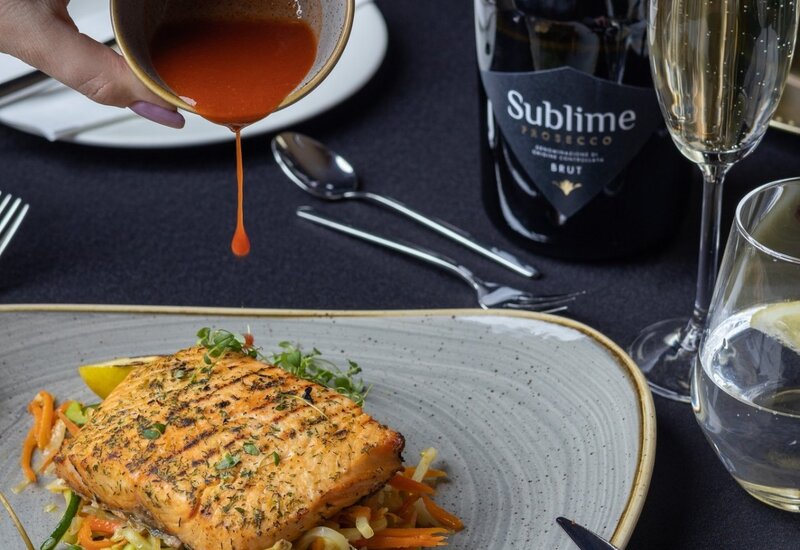 Grilled salmon on vegetables with caperberry garnish, hand pouring red sauce; bottle of Sublime Prosecco and sparkling wine flute