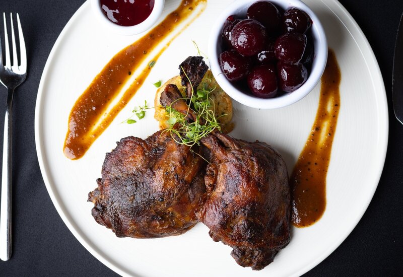 Slow-roasted duck leg with brown sauce, microgreens, potato cake and glazed cherries in a bowl on a white plate, black table setting