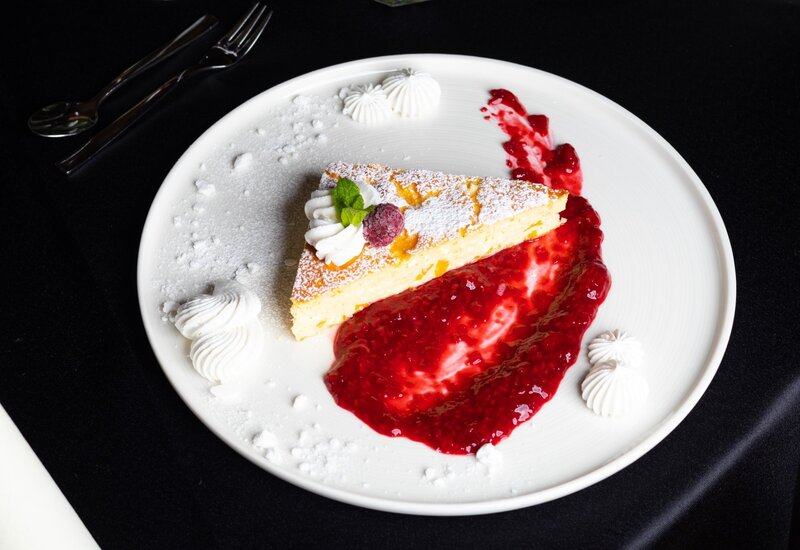 Slice of powdered-sugar cheesecake with whipped cream, raspberry and mint, served with raspberry coulis and meringue dollops on a white plate