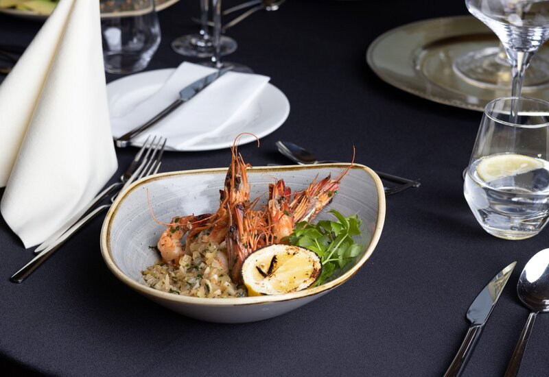 Grilled prawns with charred lemon, herbed rice and greens in a ceramic bowl on an elegant black-tablecloth table with folded napkin