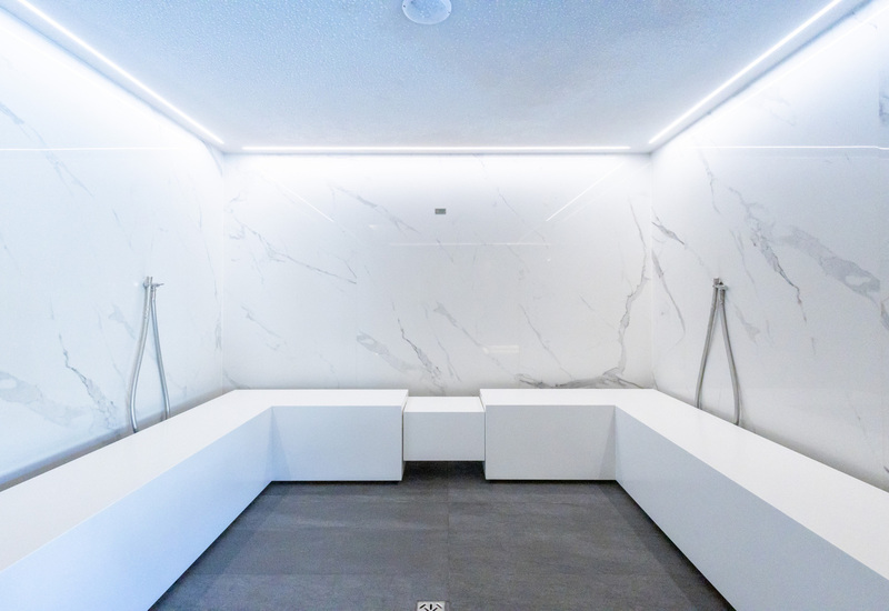 Marble-lined spa room with U-shaped white benches, two handheld shower wands and recessed LED ceiling lighting.