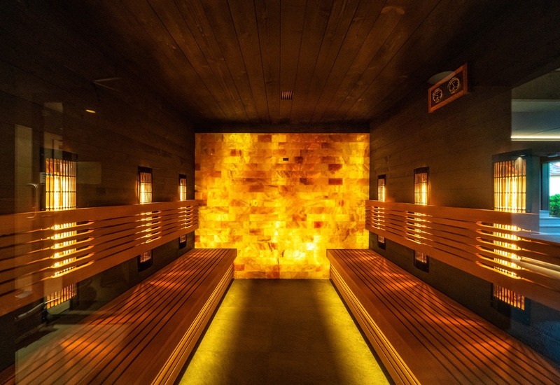 Infrared sauna with backlit Himalayan salt wall, wooden benches on both sides and warm amber lighting for relaxing spa.