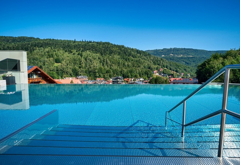 Rooftop infinity pool with steps and stainless handrail, overlooking a forested mountain valley and village under clear blue sky