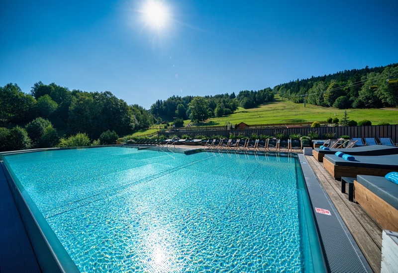 Outdoor pool with sunbeds and rolled towels on a wooden deck, view of sunlit mountain slopes, forest and chairlift