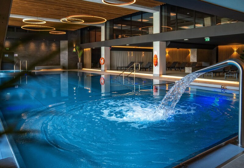 Indoor hotel pool with stainless waterfall jet, illuminated blue water, wooden slatted ceiling, circular pendant lights and poolside loungers.