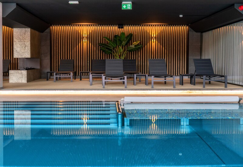 Indoor hotel pool with turquoise water, black loungers, warm wood-slat wall lighting and a large tropical plant.