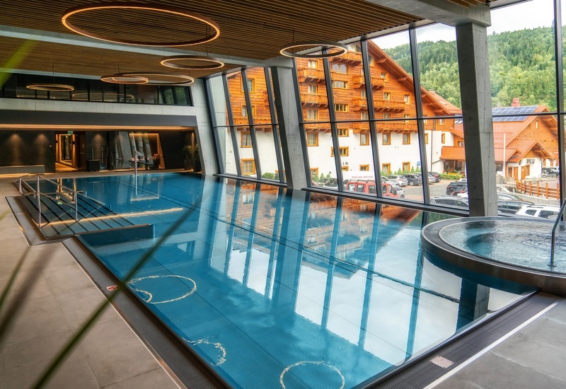 Indoor infinity-style pool with circular ceiling lights, adjacent bubbling hot tub and floor-to-ceiling windows overlooking a wooden chalet and forested mountains