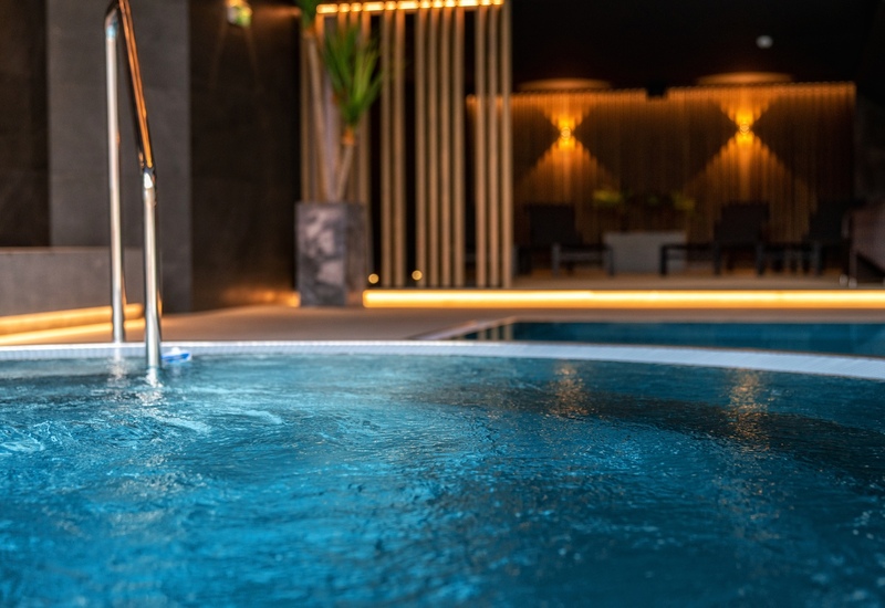 Indoor hotel spa pool with bubbling water, stainless steel handrail, warm ambient lighting and lounge chairs by a wooden slat wall