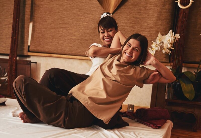 Spa therapist performing traditional stretch massage on a smiling guest in a warm treatment room with orchids and soft lighting