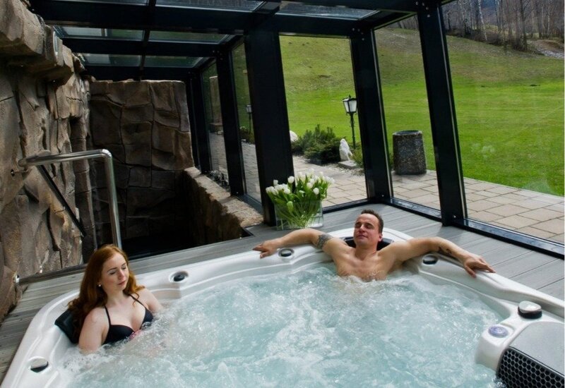 Couple relaxing in a private glass-enclosed hot tub with bubbling jets, tulips on the deck and green lawn view.