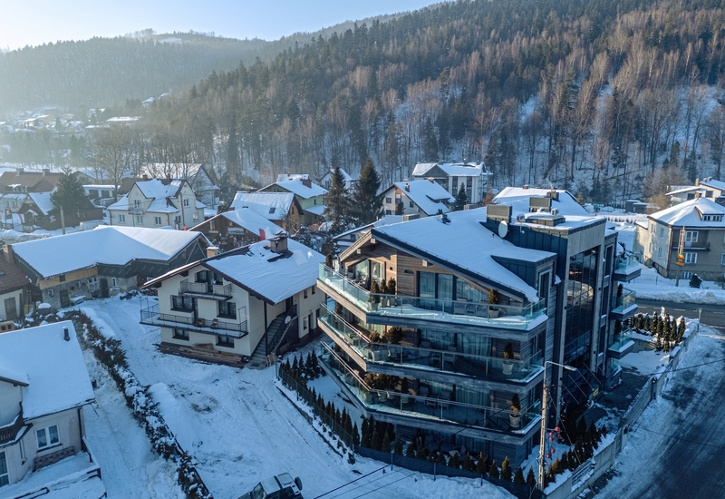 Modern glass-front hotel with wraparound balconies and terraces, snow-covered rooftops and forested mountain backdrop