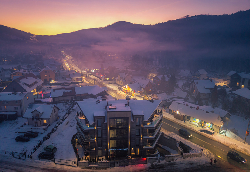 Modern hotel with glass balconies overlooking a snowy mountain village, lit shops and a misty purple‑orange sunset