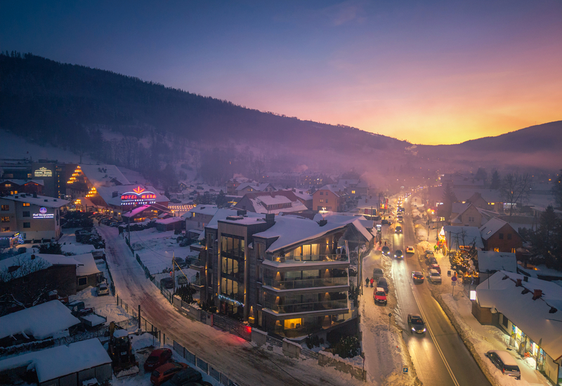 Snowy mountain resort village at sunset with illuminated hotels and spa, snow-covered roofs and a busy main street with cars and pedestrians.
