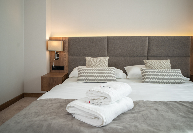 Modern twin/double hotel bed with padded headboard, wooden bedside table and lamp, patterned cushions and folded towels.