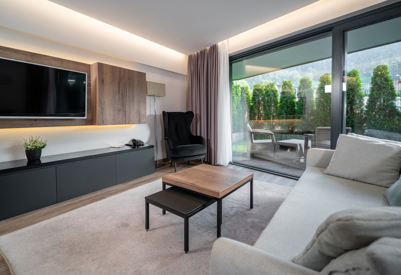 Modern suite living area with sofa and armchair, wall-mounted TV, ambient lighting and glass doors to terrace with wicker seating and mountain view.