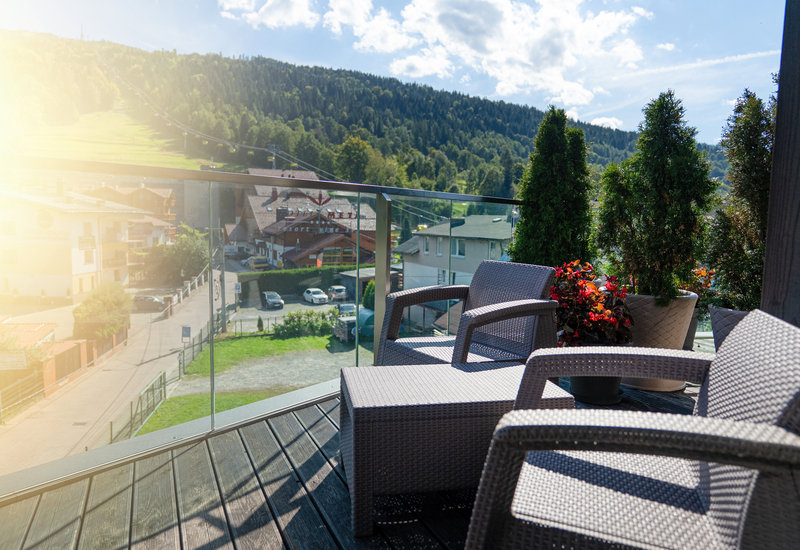 Balcony with woven lounge chairs and potted plants, glass railing overlooking village, gondola lift and forested mountain view in sunlight