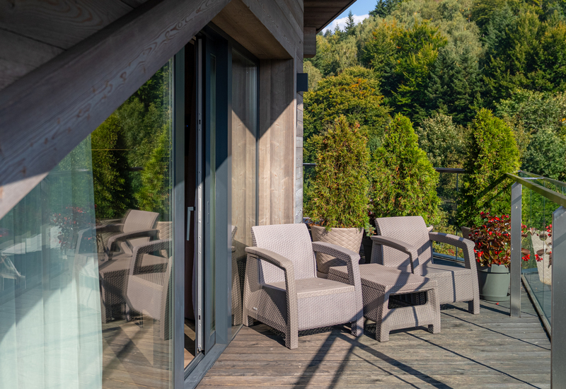 Balcony with wicker lounge chairs, potted evergreens and glass railing overlooking dense forest and mountain views