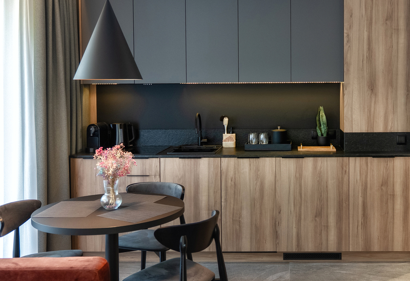Modern kitchenette in a suite with wood cabinets, black countertop, pendant light, sink, coffee machine and round dining table with vase.
