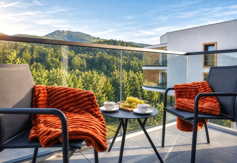 Balcony seating with orange throws, coffee cups and grapes on table, glass railing overlooking forested mountain view
