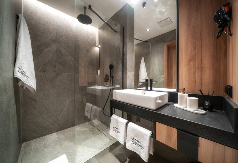 Modern hotel bathroom with walk-in rain shower, grey stone tiles, glass partition, rectangular sink on black counter, wood accents and white logo towels.