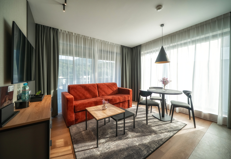 Contemporary suite living area with rust sofa, round dining table, pendant lamp, TV and floor-to-ceiling windows with hill view