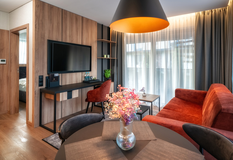 Modern hotel suite living area with red velvet sofa, wall-mounted TV, desk with bottled water, sheer curtains and balcony view.